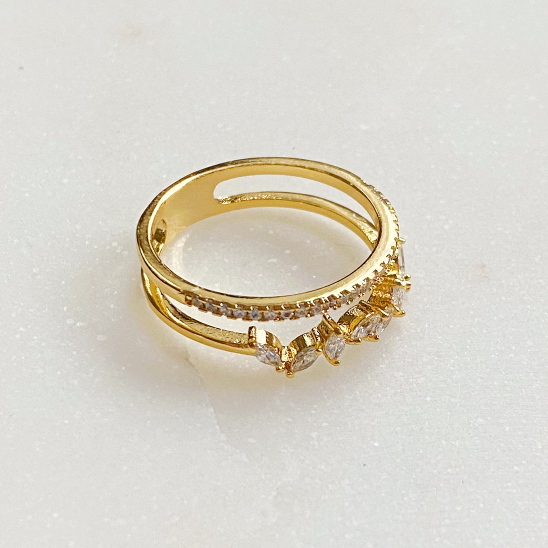 Perspective oblique d'une bague double rangée en plaqué or 18K, sertie de zircons scintillants, sur une surface marbrée, Maison Mercier.