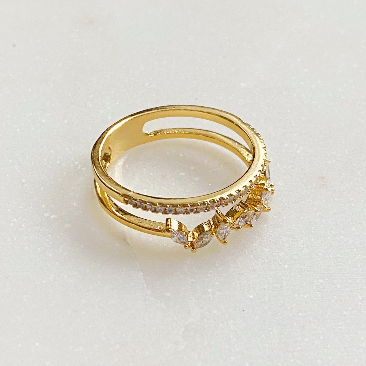 Perspective oblique d'une bague double rangée en plaqué or 18K, sertie de zircons scintillants, sur une surface marbrée, Maison Mercier.