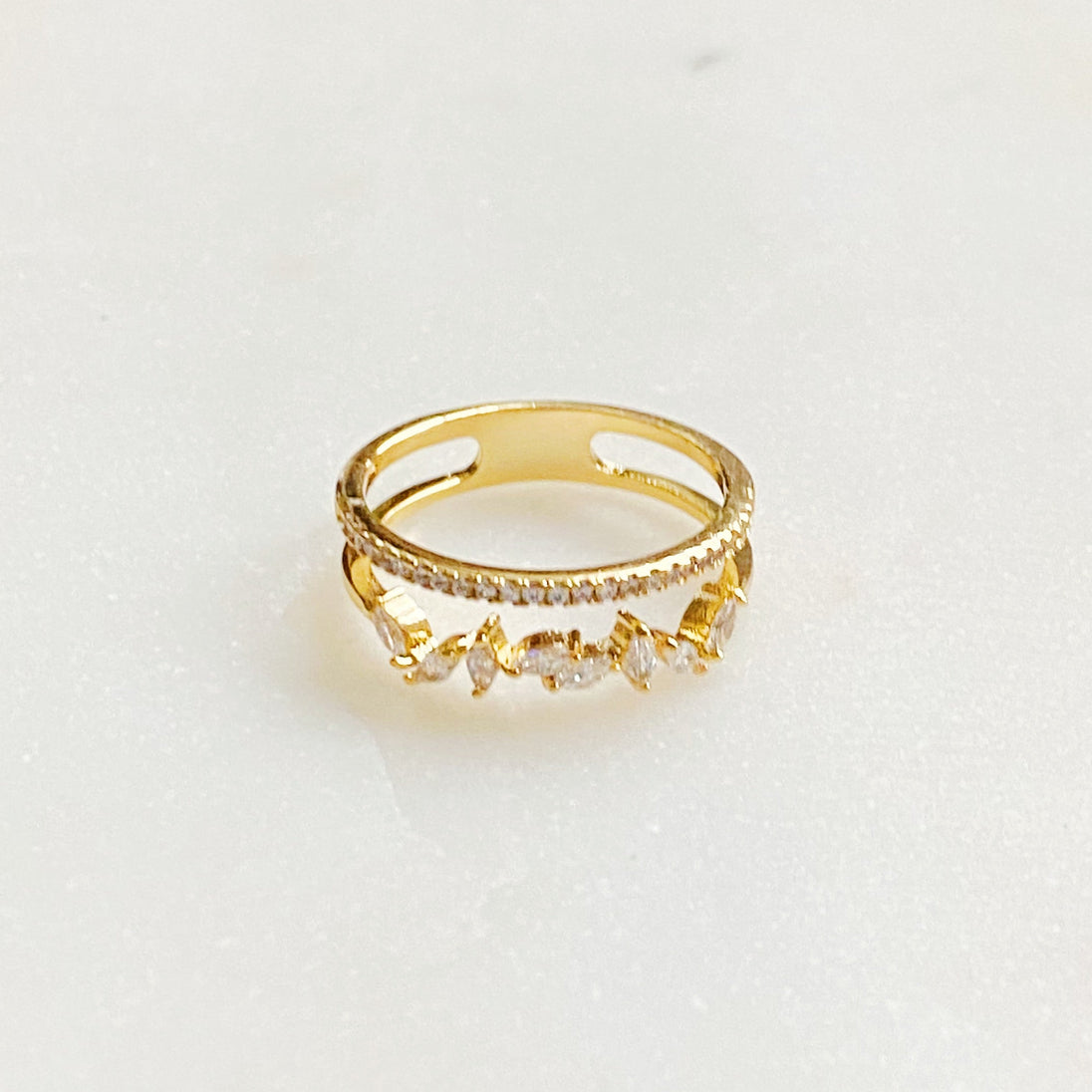 Vue de dessus d'une bague double rangée en plaqué or 18K, ornée de zircons ronds et marquises, sur un fond marbré clair, Maison Mercier.
