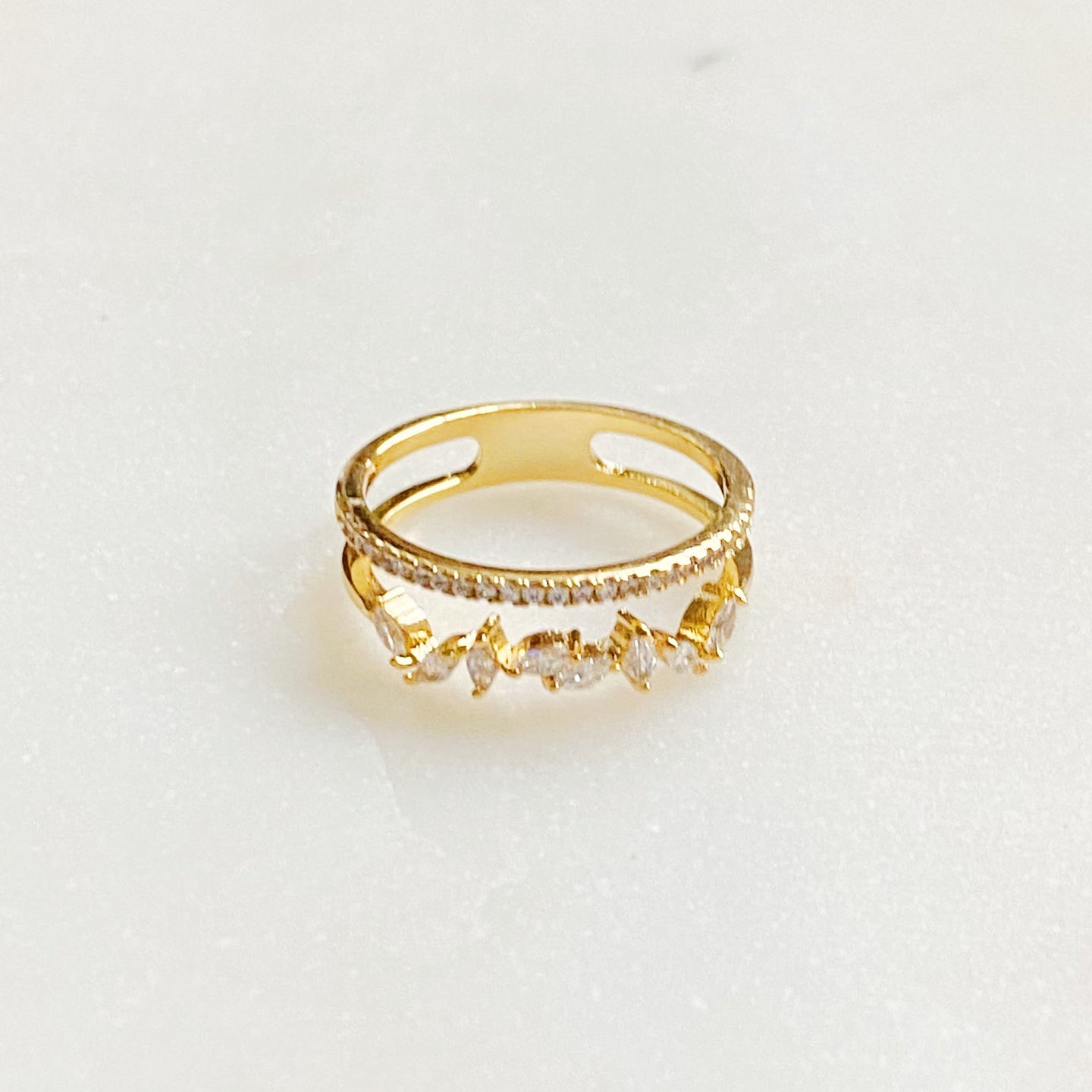 Vue de dessus d'une bague double rangée en plaqué or 18K, ornée de zircons ronds et marquises, sur un fond marbré clair, Maison Mercier.
