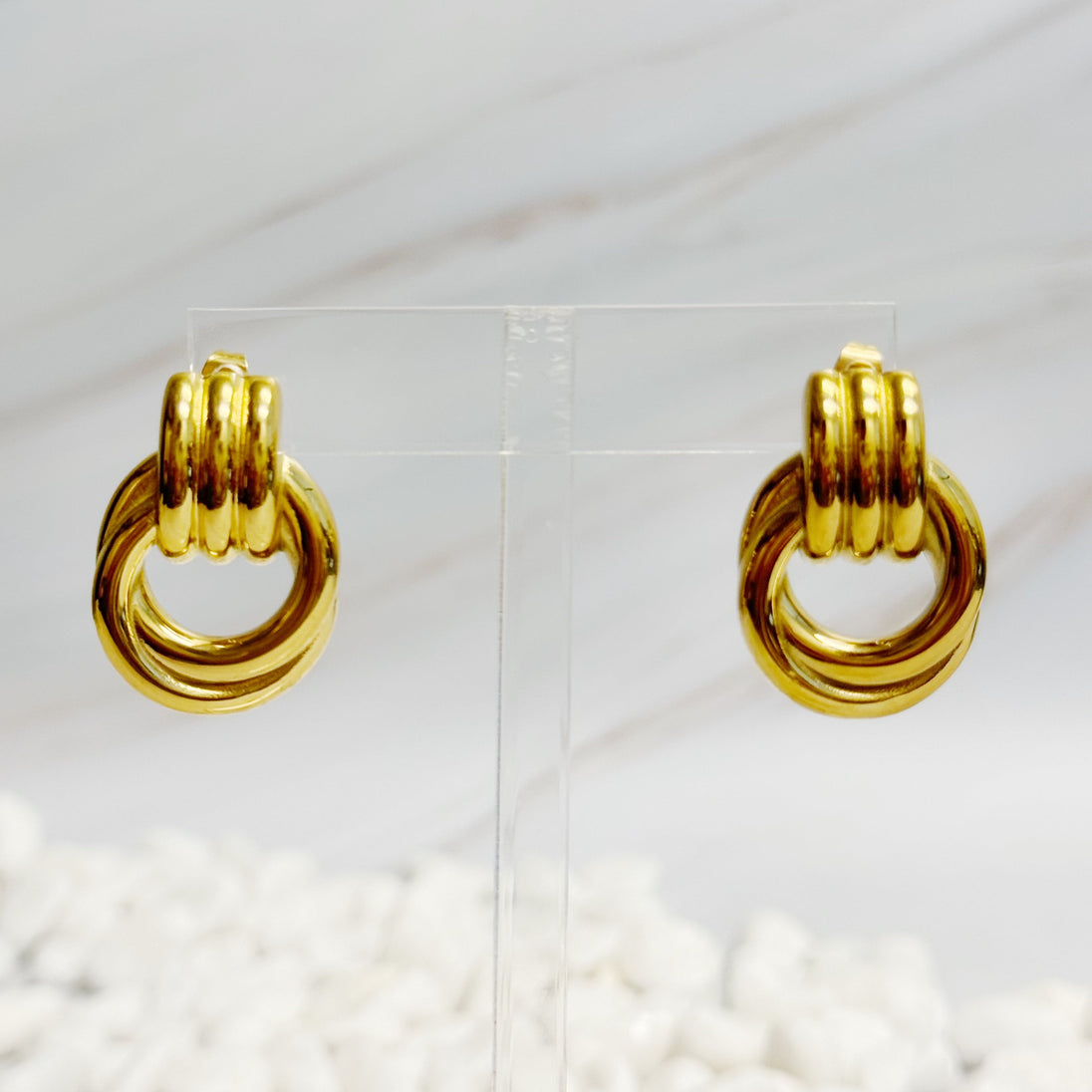 Paire de boucles d'oreilles anneaux entrelacés dorés en acier inoxydable présentées sur un support transparent, Maison Mercier.