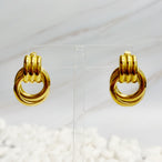 Paire de boucles d'oreilles anneaux entrelacés dorés en acier inoxydable présentées sur un support transparent, Maison Mercier.