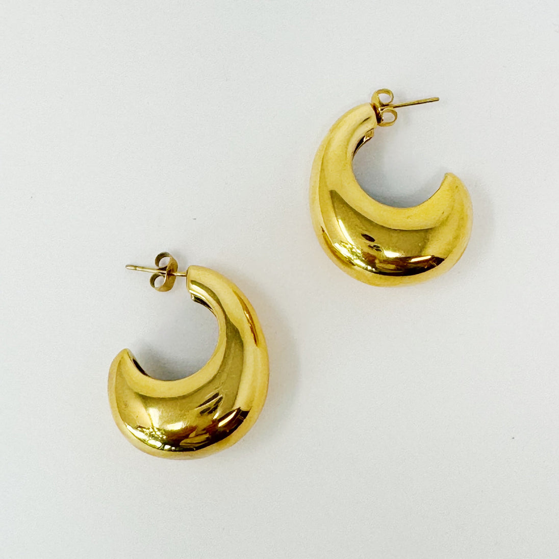 Détail d'une paire de boucles d'oreilles créoles bombées en acier doré poli, reflétant la lumière sur fond blanc.