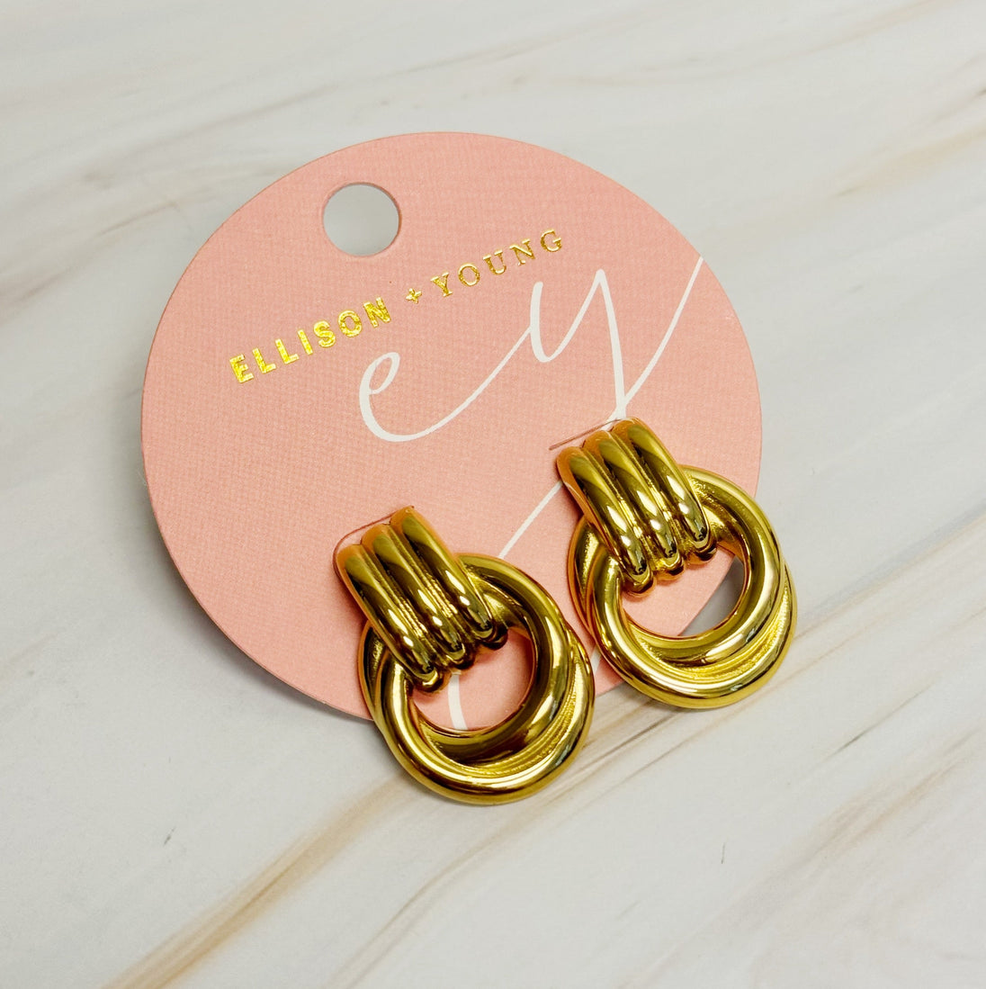 Détail de deux boucles d'oreilles anneaux entrelacés dorés en acier inoxydable poli sur un fond de marbre clair, Maison Mercier.