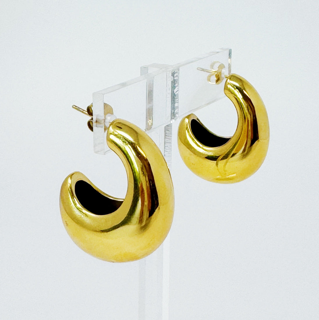 Boucles d'oreilles créoles bombées en acier inoxydable doré, présentées sur un support transparent, illustrant leur volume élégant.