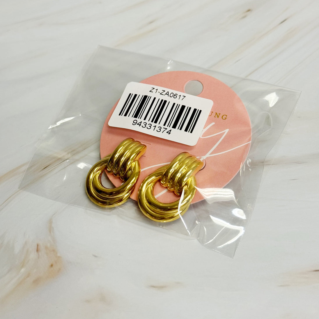Détail de deux boucles d'oreilles anneaux entrelacés dorés en acier inoxydable poli sur un fond de marbre clair, Maison Mercier.