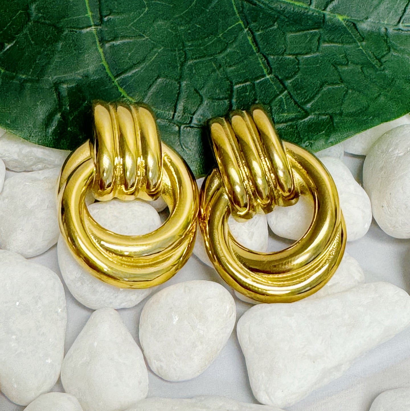 Vue rapprochée de deux boucles d'oreilles anneaux entrelacés dorés en acier inoxydable sur un lit de galets blancs avec une feuille verte, Maison Mercier.