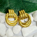 Vue rapprochée de deux boucles d'oreilles anneaux entrelacés dorés en acier inoxydable sur un lit de galets blancs avec une feuille verte, Maison Mercier.