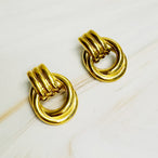 Détail de deux boucles d'oreilles anneaux entrelacés dorés en acier inoxydable poli sur un fond de marbre clair, Maison Mercier.