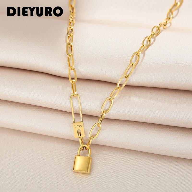 Collier gourmette ovale doré avec cadenas et épingle 'Love' en acier inoxydable, logo DIEURO en haut à gauche, sur un tissu soyeux beige, Maison Mercier.