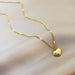 Détail d'un collier pendentif cœur 'Love' en acier inoxydable doré poli, posé sur un tissu élégant, Maison Mercier.