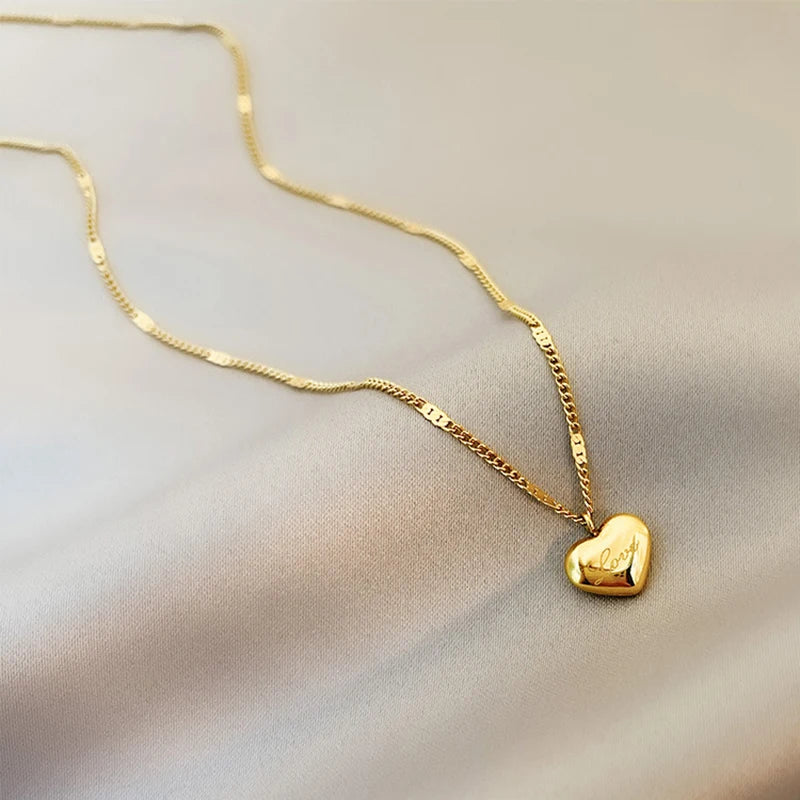 Détail d'un collier pendentif cœur 'Love' en acier inoxydable doré poli, posé sur un tissu élégant, Maison Mercier.