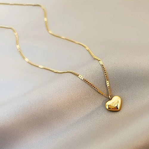 Détail d'un collier pendentif cœur 'Love' en acier inoxydable doré poli, posé sur un tissu élégant, Maison Mercier.