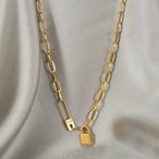 Collier gourmette ovale doré avec cadenas et épingle 'Love' en acier inoxydable, posé sur un tissu soyeux gris clair, Maison Mercier.