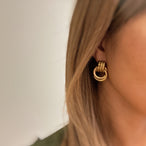 Détail de deux boucles d'oreilles anneaux entrelacés dorés en acier inoxydable poli sur un fond de marbre clair, Maison Mercier.