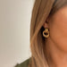 Détail de deux boucles d'oreilles anneaux entrelacés dorés en acier inoxydable poli sur un fond de marbre clair, Maison Mercier.