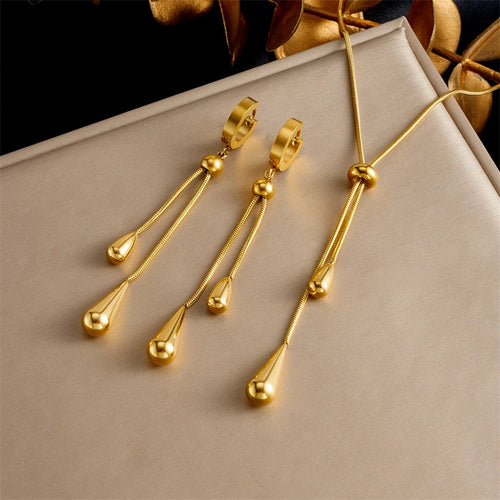 Parure Goutte d'Eau en acier doré, collier lariat et boucles pendantes, présentée sur un fond élégant et texturé, Maison Mercier.