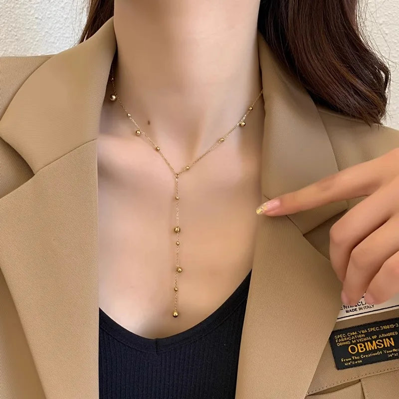 Collier Lariat Y en acier doré, avec ses perles polies, reflétant la lumière sur une surface claire.