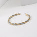 Bracelet jonc ouvert torsadé doré en acier inoxydable, présenté dans un écrin de velours rouge, avec une étiquette Saint Laurent et une carte Joyful Nails Studio.