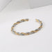 Bracelet jonc ouvert torsadé doré en acier inoxydable, présenté dans un écrin de velours rouge, avec une étiquette Saint Laurent et une carte Joyful Nails Studio.