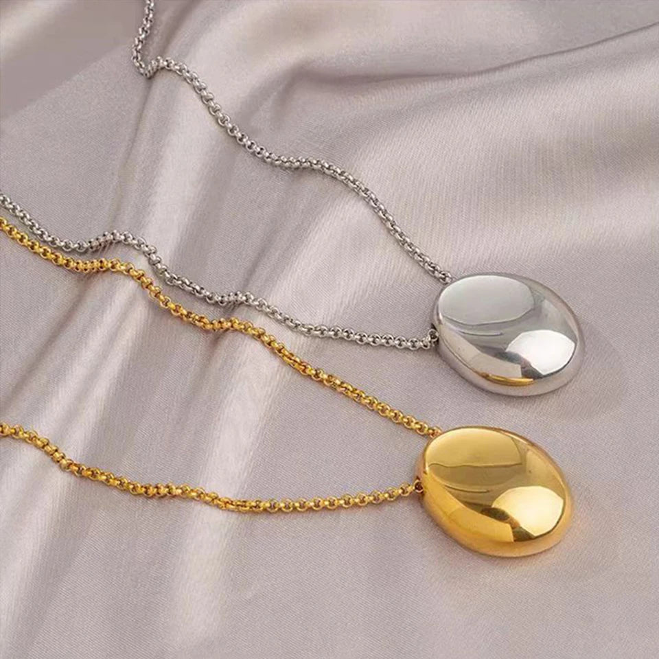 Deux colliers pendentifs ovales sculpturaux polis, l'un en finition argentée et l'autre en finition dorée, posés sur un tissu soyeux, Maison Mercier.