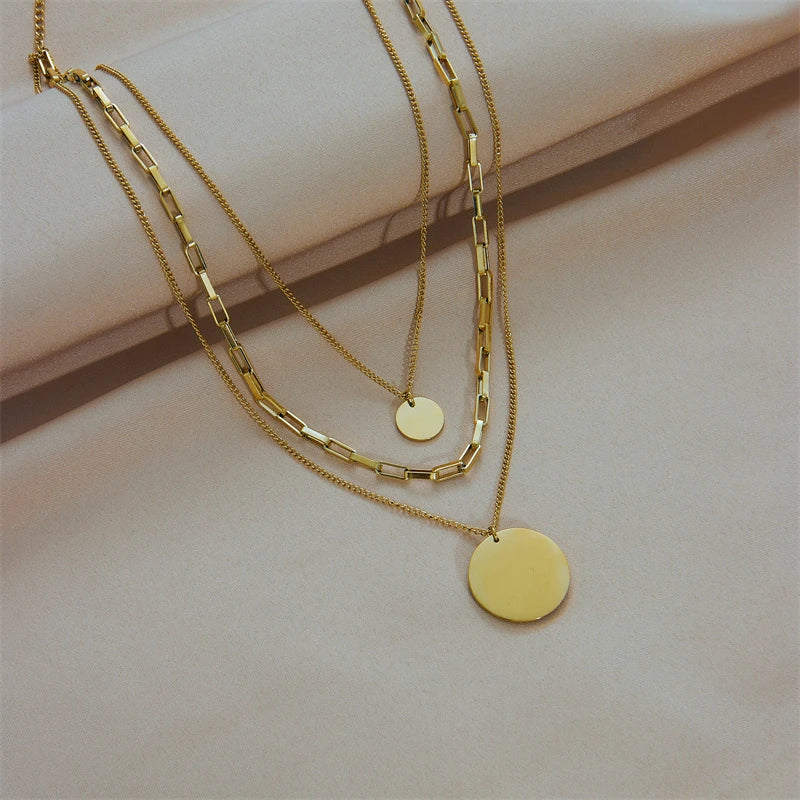 Collier multirangs doré avec chaînes fines et maillons architecturaux, et pendentifs disques polis, posé sur un tissu soyeux beige, Maison Mercier.