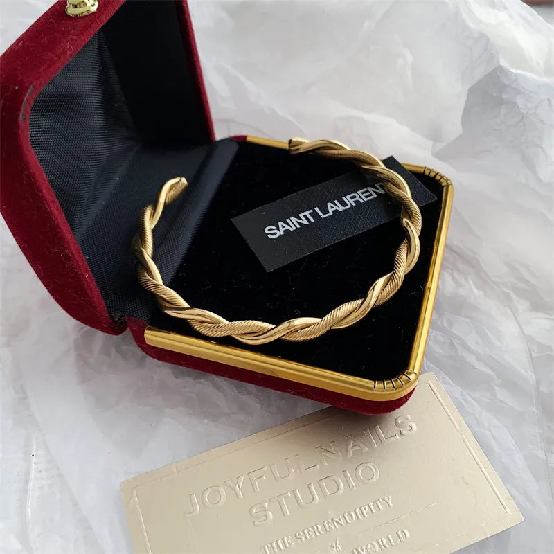 Bracelet jonc ouvert torsadé doré en acier inoxydable, présenté dans un écrin de velours rouge, avec une étiquette Saint Laurent et une carte Joyful Nails Studio.