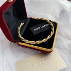 Bracelet jonc ouvert torsadé doré en acier inoxydable, présenté dans un écrin de velours rouge, avec une étiquette Saint Laurent et une carte Joyful Nails Studio.