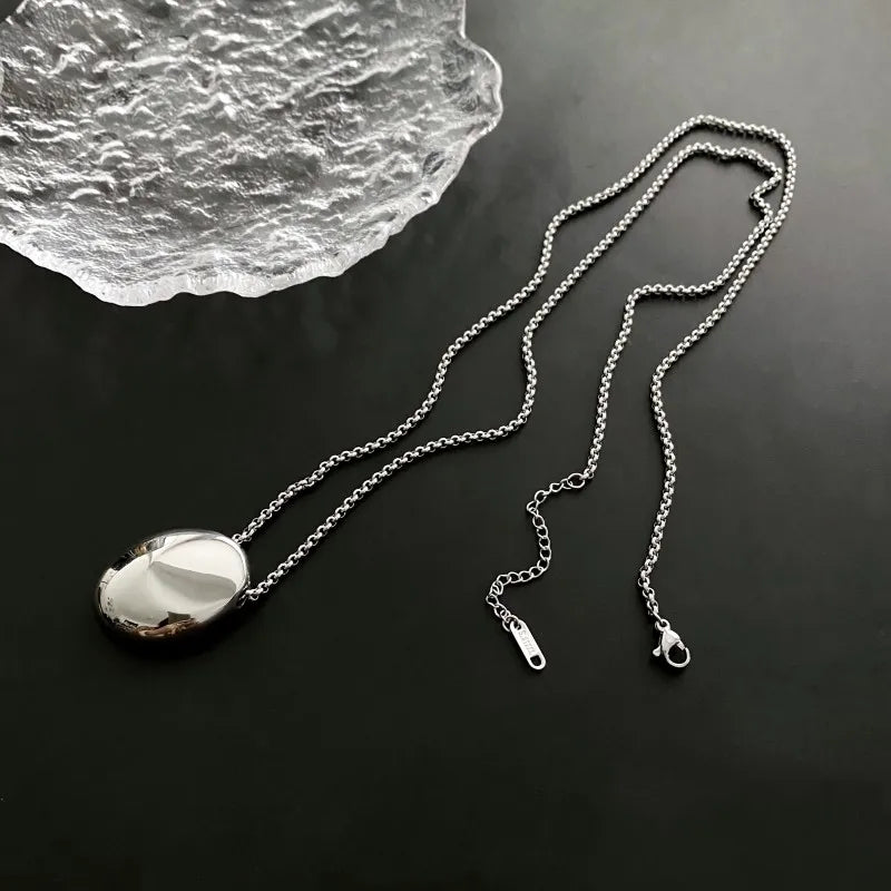 Collier pendentif ovale sculptural poli en finition argentée, posé sur une surface sombre à côté d'un élément décoratif en verre texturé, Maison Mercier.