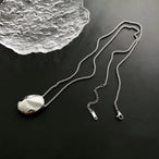 Collier pendentif ovale sculptural poli en finition argentée, posé sur une surface sombre à côté d'un élément décoratif en verre texturé, Maison Mercier.