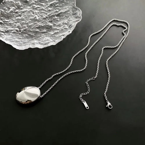 Deux colliers pendentifs ovales sculpturaux polis, l'un en finition argentée et l'autre en finition dorée, posés sur un tissu soyeux, Maison Mercier.