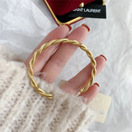 Main portant un pull en maille crème, présentant un bracelet jonc ouvert torsadé doré en acier inoxydable, mettant en valeur sa texture et sa finition polie.