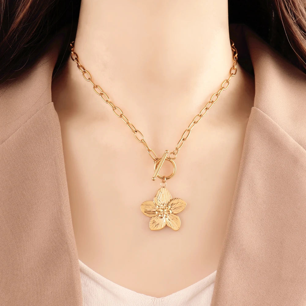 Collier chaîne ovale en acier inoxydable doré, avec pendentif fleur sculptée et fermoir T-Bar, présenté sur un écrin beige satiné, Maison Mercier.