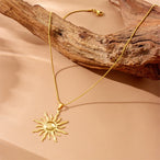Collier pendentif soleil rayonnant en acier inoxydable doré, posé délicatement sur une pièce de bois flotté, Maison Mercier.