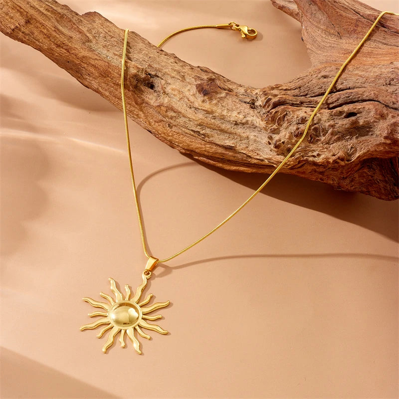Collier pendentif soleil rayonnant en acier inoxydable doré, posé délicatement sur une pièce de bois flotté, Maison Mercier.