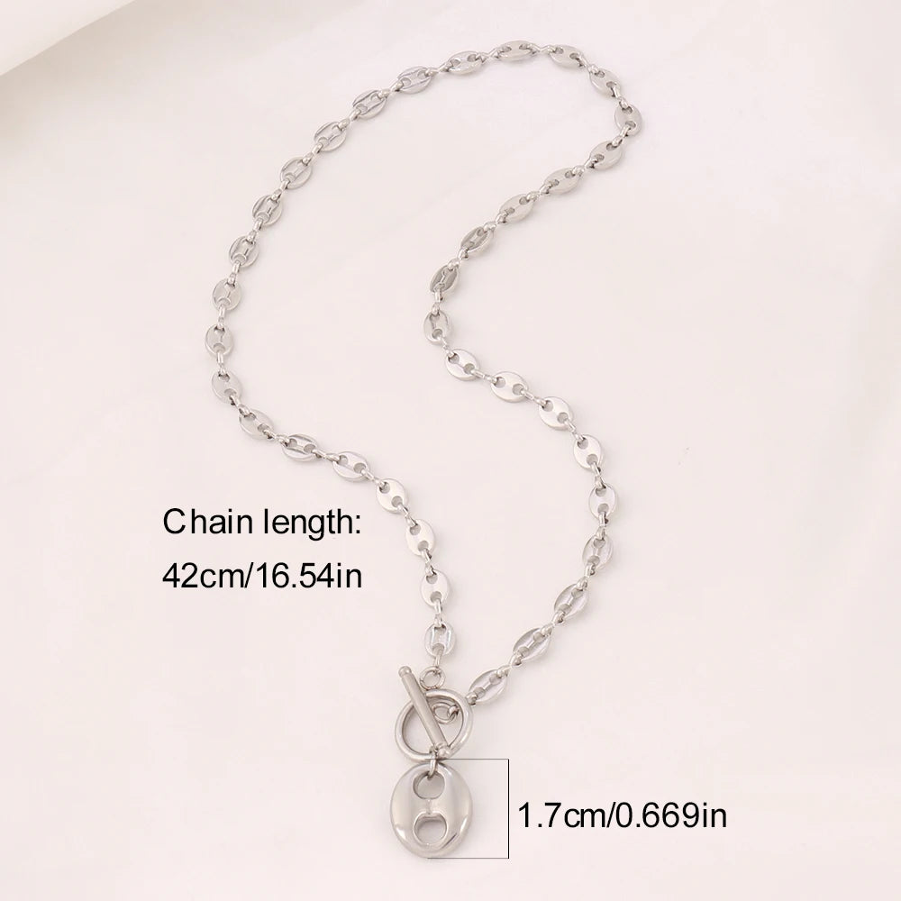 Collier Grains de Café en acier inoxydable doré, fermoir toggle, posé sur un tissu blanc et une pierre texturée, Maison Mercier.