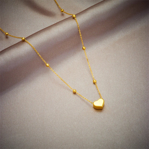 Détail d'un collier pendentif cœur 'Love' en acier inoxydable doré poli, posé sur un tissu élégant, Maison Mercier.