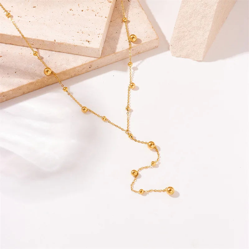 Gros plan sur la chute du collier Lariat Y doré, avec ses sphères polies, posé sur des blocs de pierre clairs.