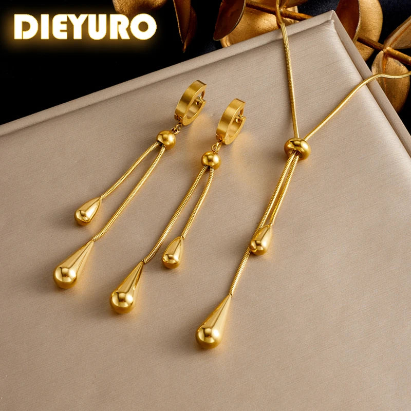 Parure Goutte d'Eau en acier doré, collier lariat et boucles pendantes, présentée sur un fond élégant et texturé, Maison Mercier.