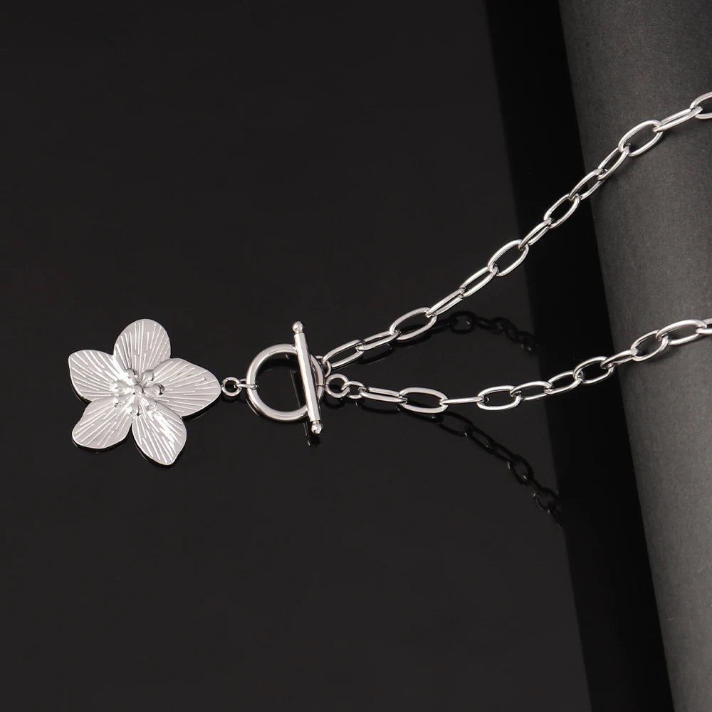 Collier chaîne ovale en acier inoxydable argenté, avec pendentif fleur sculptée et fermoir T-Bar, élégamment posé sur un fond noir, Maison Mercier.