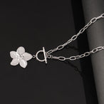 Collier chaîne ovale en acier inoxydable argenté, avec pendentif fleur sculptée et fermoir T-Bar, élégamment posé sur un fond noir, Maison Mercier.