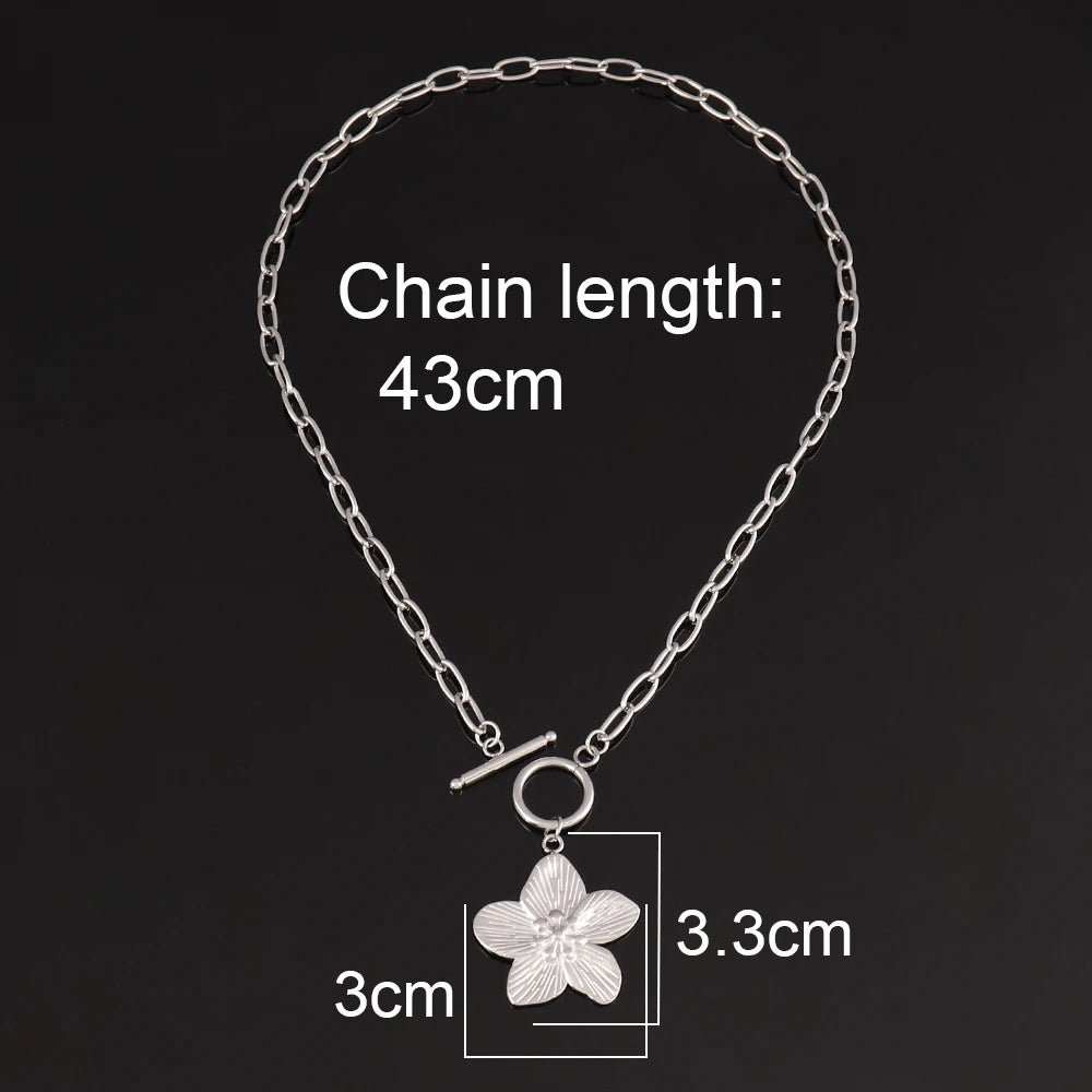 Collier chaîne ovale en acier inoxydable doré, avec pendentif fleur sculptée et fermoir T-Bar, présenté sur un écrin beige satiné, Maison Mercier.