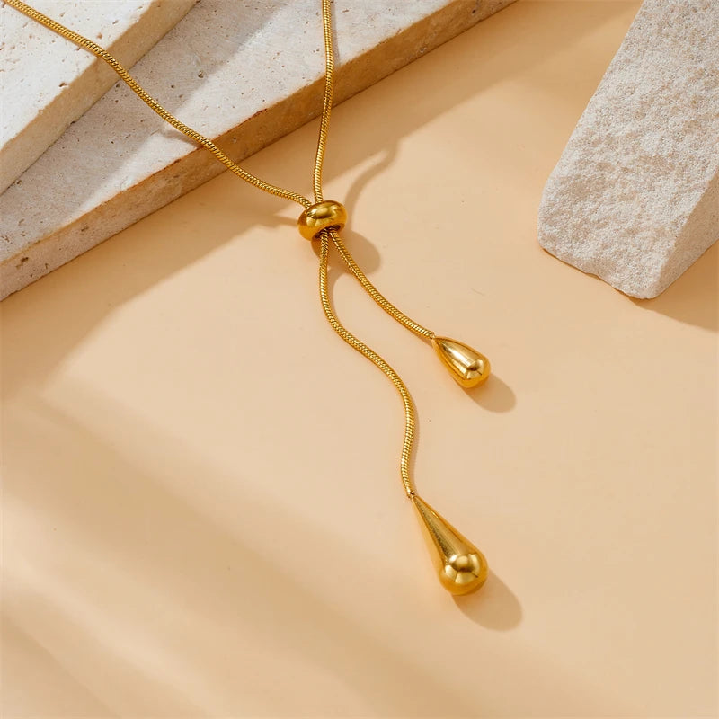 Parure Goutte d'Eau en acier doré, collier lariat et boucles pendantes, présentée sur un fond élégant et texturé, Maison Mercier.
