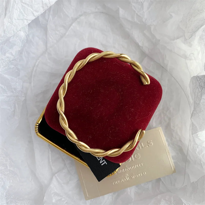 Vue de dessus d'un bracelet jonc ouvert torsadé doré en acier inoxydable, posé sur un écrin de velours rouge, avec une carte dorée en arrière-plan.