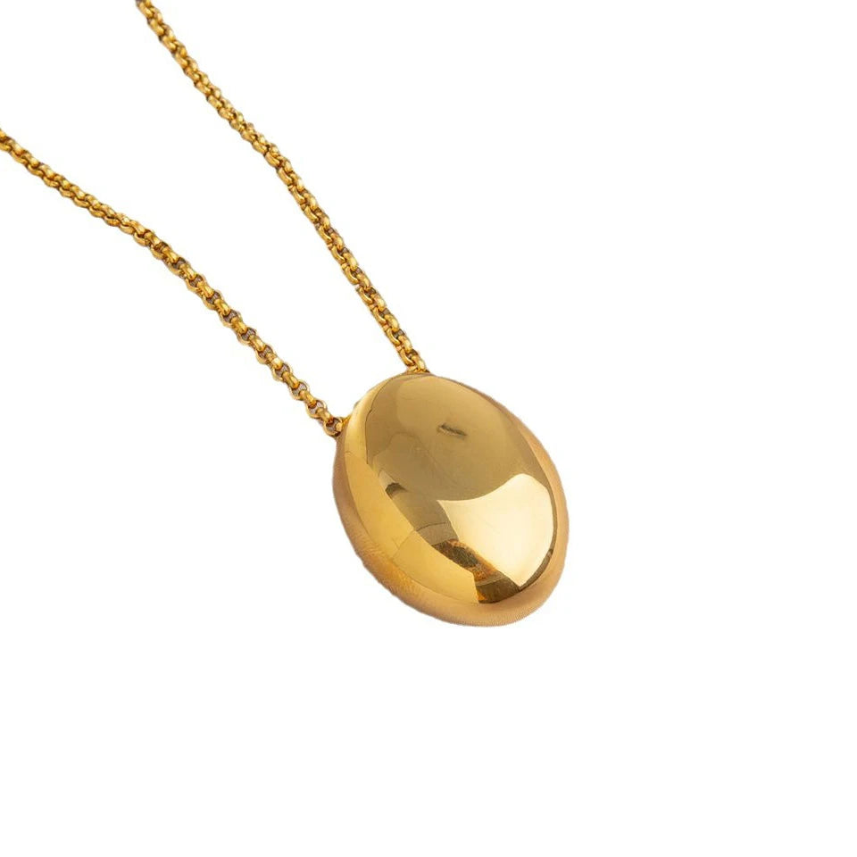 Deux colliers pendentifs ovales sculpturaux polis, l'un en finition argentée et l'autre en finition dorée, posés sur un tissu soyeux, Maison Mercier.