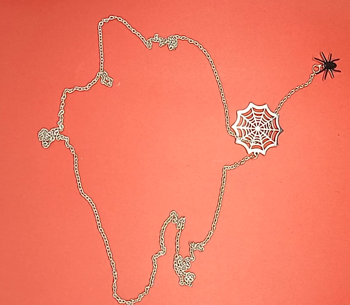Collier pendentif toile d'araignée et araignée en finition argentée, la chaîne délicatement posée sur un fond corail, Maison Mercier.