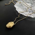 Deux colliers pendentifs ovales sculpturaux polis, l'un en finition argentée et l'autre en finition dorée, posés sur un tissu soyeux, Maison Mercier.
