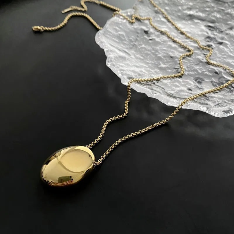 Deux colliers pendentifs ovales sculpturaux polis, l'un en finition argentée et l'autre en finition dorée, posés sur un tissu soyeux, Maison Mercier.