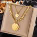 Collier multirangs en acier inoxydable doré avec pendentifs disques polis, présenté sur un écrin beige, Maison Mercier.