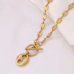 Collier Grains de Café en acier inoxydable doré, fermoir toggle, posé sur un tissu blanc et une pierre texturée, Maison Mercier.
